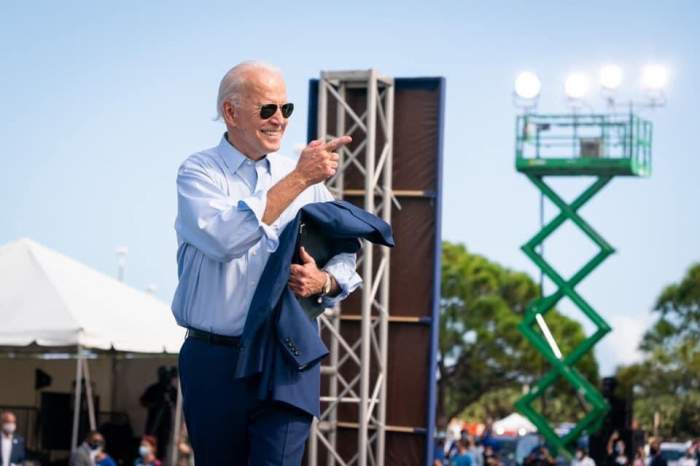 Joe Biden se află pe scenă. Acesta își ține în mână sacoul, e îmbrăcat cu o cămașă bleu și pantaloni albaștri, iar pe ochi are potriviți ochelarii de soare