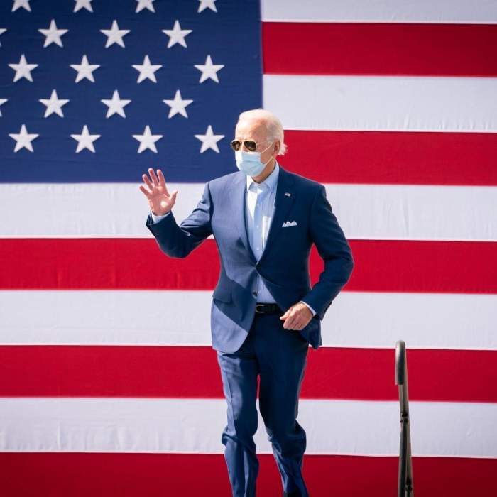 Joe Biden poartă mască de protecție și un costum albastru. Acesta le face cu mâna oamenilor.