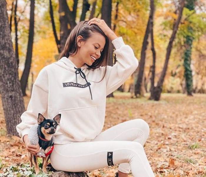 Denisa Hodișan e îmbrăcată într-un trening alb. Vedeta se află în parc, stă pe o buturugă, iar lângă ea se află câinele ei.