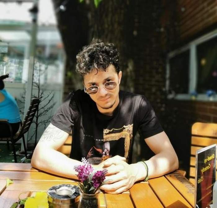 Uddi se află la terasă. Artistul poartă ochelari de soare și un tricou negru cu imprimeu și ține mâinile pe masă.