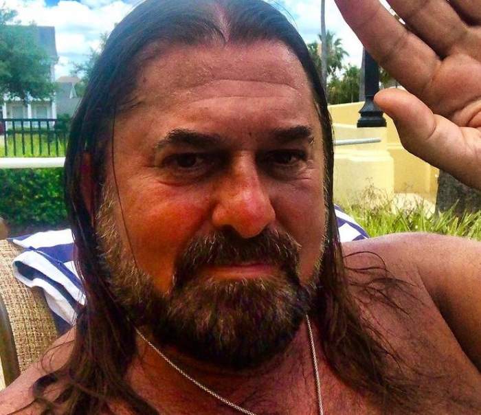 Gheorghe Gheorghiu își face un selfie și ține o mână ridicată, în semn de salut. Artistul nu poartă tricou și are un lanț argintiu la gât.