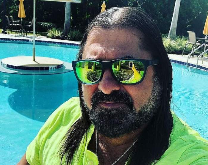 Gheorghe Gheorghiu se află la piscină. Aritstul poartă o pereche de ochelari de soare și un tricou verde.