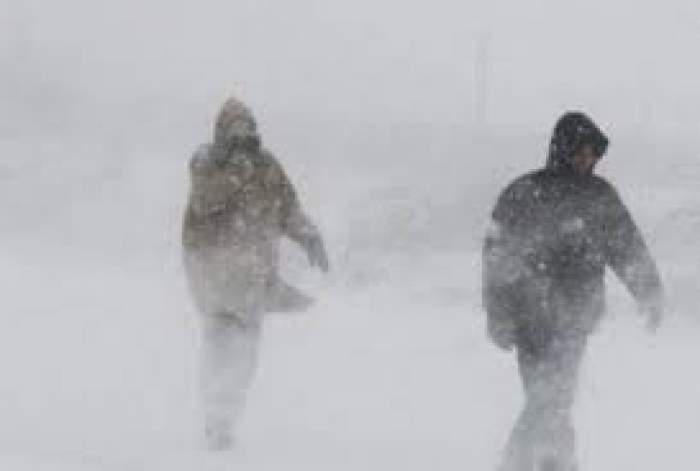 Doi oameni, îmbrăcați de iarnă, merg prin vânt și ninsori