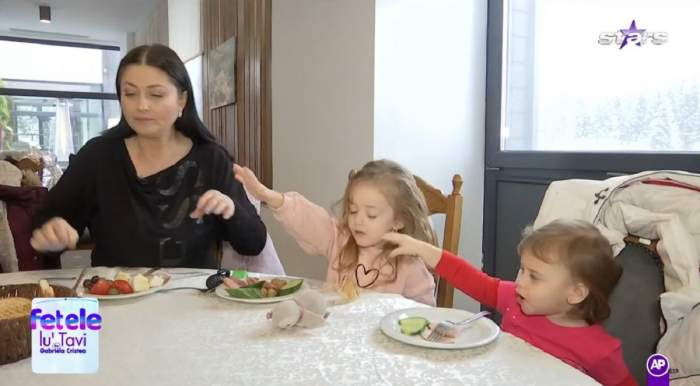Gabriela Cristea și fetițele ei iau micul-dejun. Vedeta de la Antena Stars poartă o bluză neagră, Victoria una roz și Iris una roșie. Toate trei au în față platouri cu mâncare.