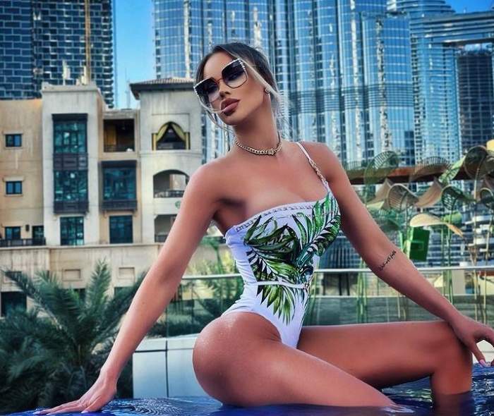 Bianca Drăgușanu se află în Dubai și e așezată pe marginea piscinei. Vedeta poartă un costum de baie alb cu model cu frunze verzi și are părul prins în coc.