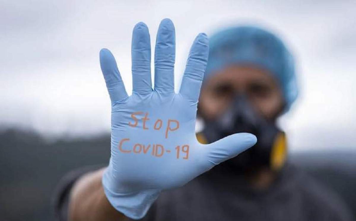O persoană care poartă mănuși ține mâna întinsă în față, iar în palmă are scris: stop covid-19.