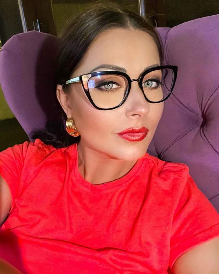 Gabriela Cristea stă pe un fotoliu mov. Vedeta poartă un tricou roșu și ochelari de vedere.