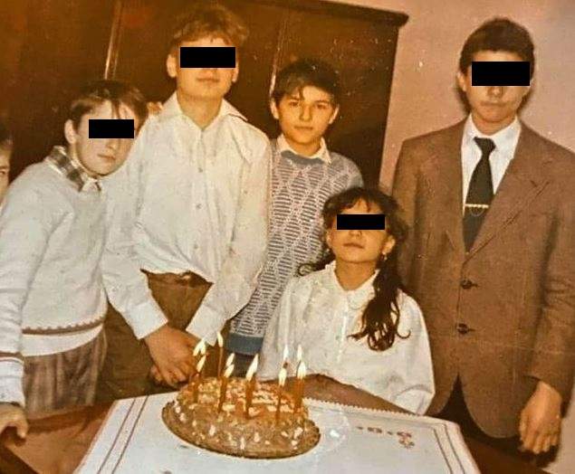 Liviu Vârciu în copilărie. Artistul purta o cămașă albă, iar pe deasupra un pulover cu nuanțe de gri. Lângă el sunt mai mulți copii, iar în fața lor o masă cu un tort.