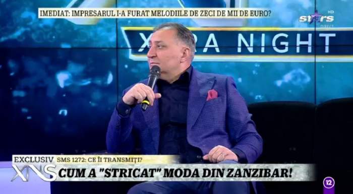 Vali Vijelie se află la Xtra Night Show și stă pe canapea. Artistul poartă un costum albastru.