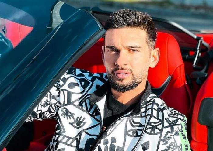 Dorian Popa se află într-o mașină decapotabilă cu tapițerie roșie. Artistul poartă un hanorac alb cu model negru.