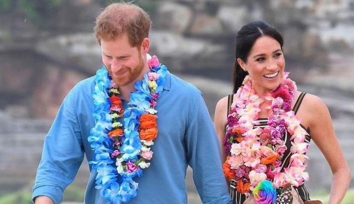 Prințul Harry și Meghan Markle zâmbesc. Ea poartă o rochie neagră, iar el o cămașă bleu.