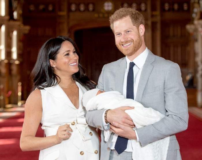 Meghan Markle poartă o rochie albă, tip sacou și este alături de soțul ei. Prințul Harry îl ține pe fiul lor în brațe.