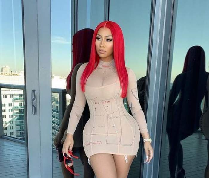 Nicki Minaj poartă o rochie mulată de culoare crem. Vedeta are în mână o pereche de ochelari de soare roșii și are părul în aceeași nuanță.