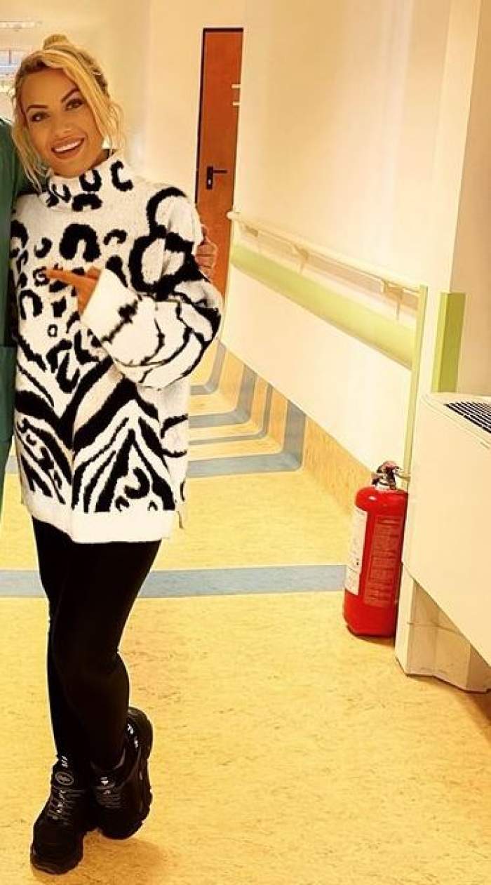 Amna se află la spital. Artista zâmbește larg, poartă un pulover cu animal print în alb și negru, pantaloni și bocanci negri.