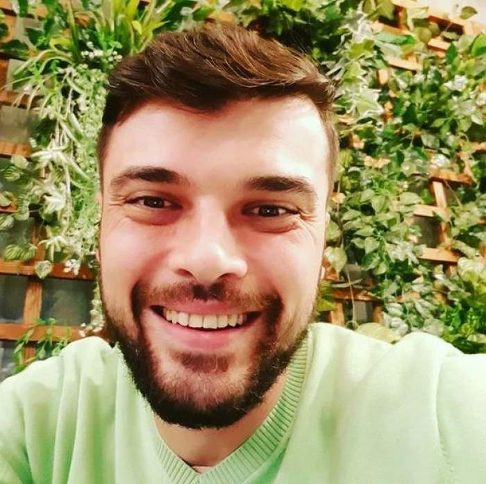 Marius Elisei într-un selfie. Bărbatul poartă un pulover verde și zâmbește. În spatele lui e un gărduleț din lemn cu plante verzi.