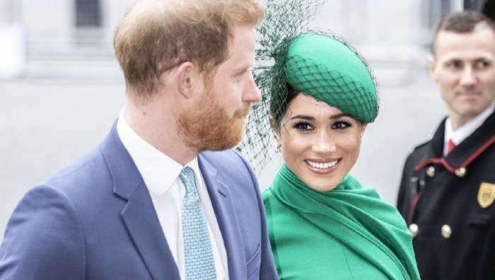 Prințul Harry și Meghan Markle se află pe stradă. Ea poartă un costum și o pălărie verde, iar el un costum bleu, cu cămașă albă. Ducesa de Sussex zâmbește larg.
