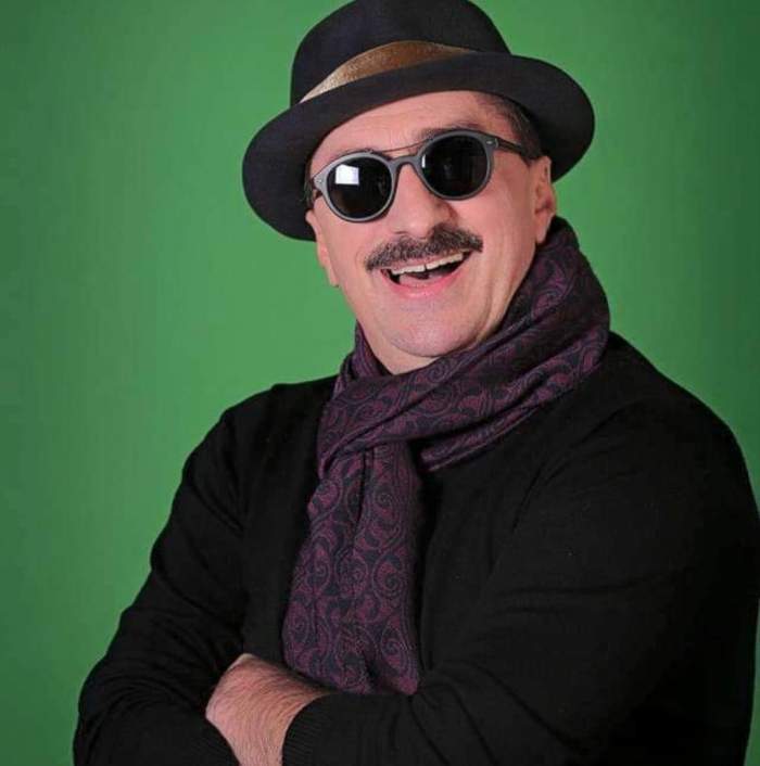 Romică Țociu poartă o pălărie neagră, ochelari de soare și o bluză în aceeași nuanță. Actorul are o eșarfă mov la gât și zâmbește.