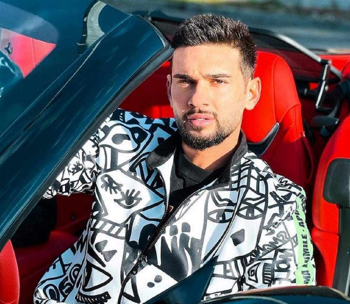 Dorian Popa se află într-o mașină decapotabilă cu tapițerie roșie. Artistul poartă un hanorac alb cu model negru.