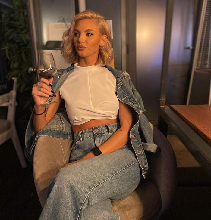 Ramona Olaru stă pe scaun, cu un pahar de vin în mână, în blugi și tricou alb