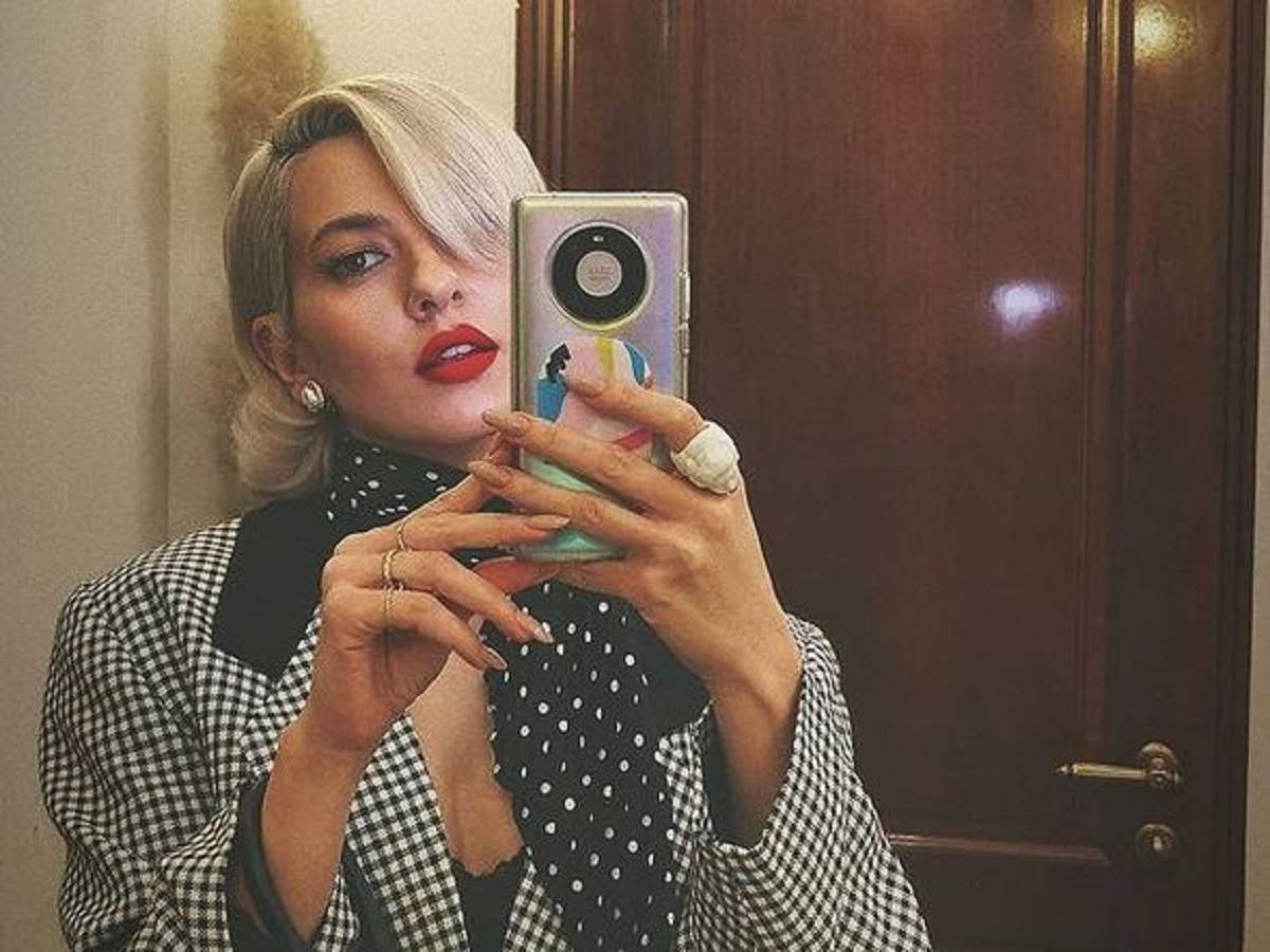 Lidia Buble își face o poză cu telefonul în oglindă. Vedeta poartă eșarfă neagră cu buline albe și sacou în pătrățele albe și negre.