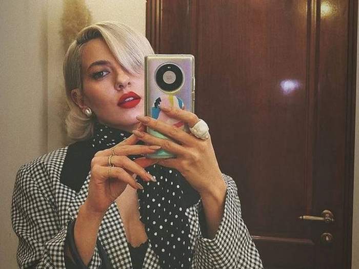 Lidia Buble își face o poză cu telefonul în oglindă. Vedeta poartă eșarfă neagră cu buline albe și sacou în pătrățele albe și negre.