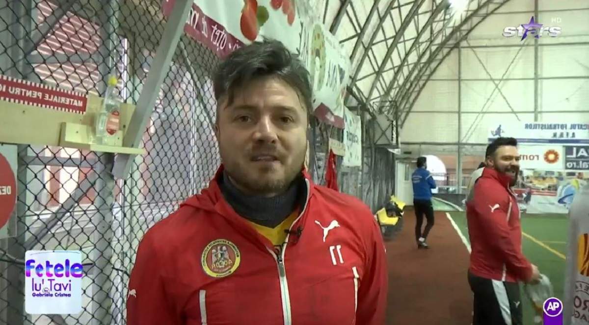 Tavi Clonda se află pe terenul de fotbal. Artistul poartă o bluză de trening roșie. În spatele lui se văd mai mulți jucători.