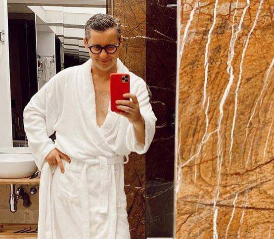 Adi Sînă poartă un halat alb și ochelari de vedere. Artistul își face o poză cu telefonul mobil în oglindă și se află în baie.