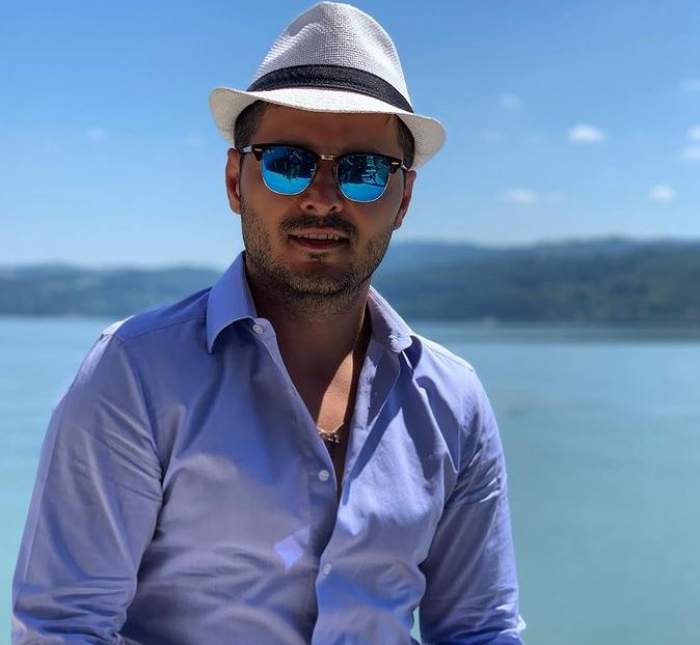 Liviu Vârciu se află la mare. Artistul poartă o pereche de ochelari de soare, o pălărie albă cu dungă neagră și o cămașă bleu.