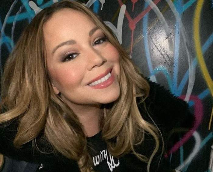 Mariah Carey își face un selfie. Vedeta poartă o geacă de iarnă neagră și zâmbește larg.