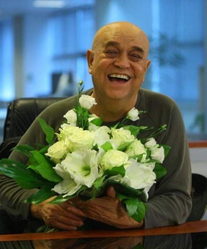 Benone Sinulescu ține în mână un buchet de flori albe. Artistul zâmbește larg.
