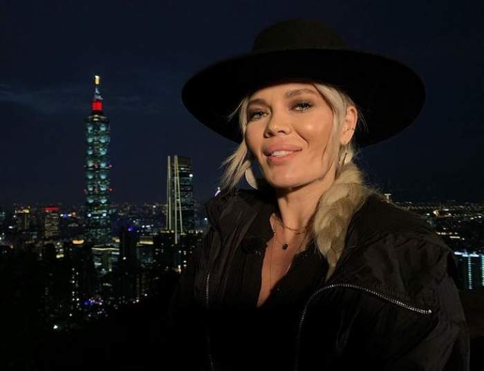 Gina Pistol poartă o geacă și pălărie neagră. În spatele ei se pot vedea luminile orașului.