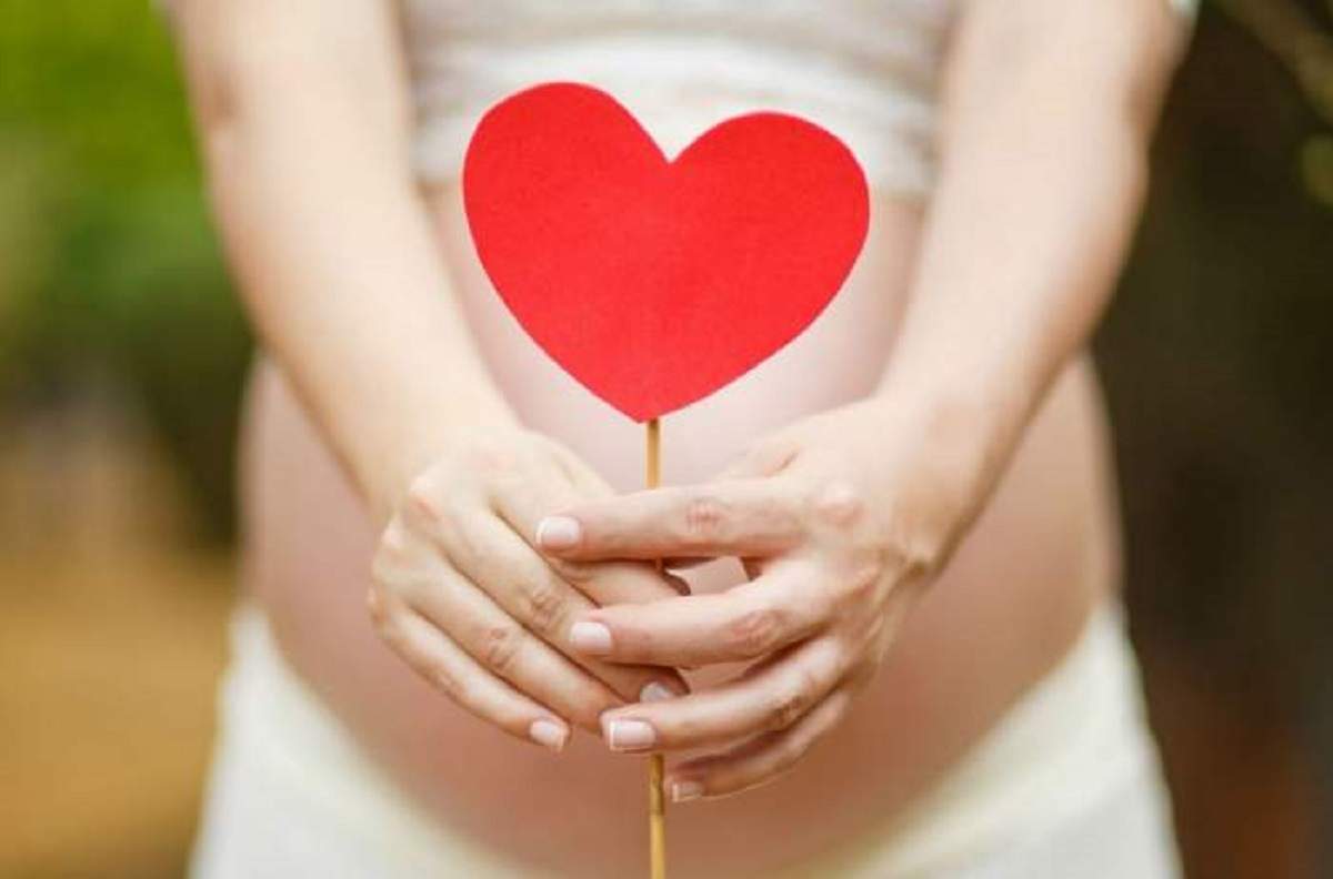 O femeie însărcinată ține o inimoară de hârtie în mână. Aceasta poartă fustă albă.