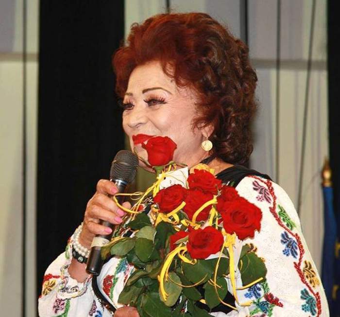 Maria Ciobanu canta la microfon, are costum popular si un buchet de flori in mana