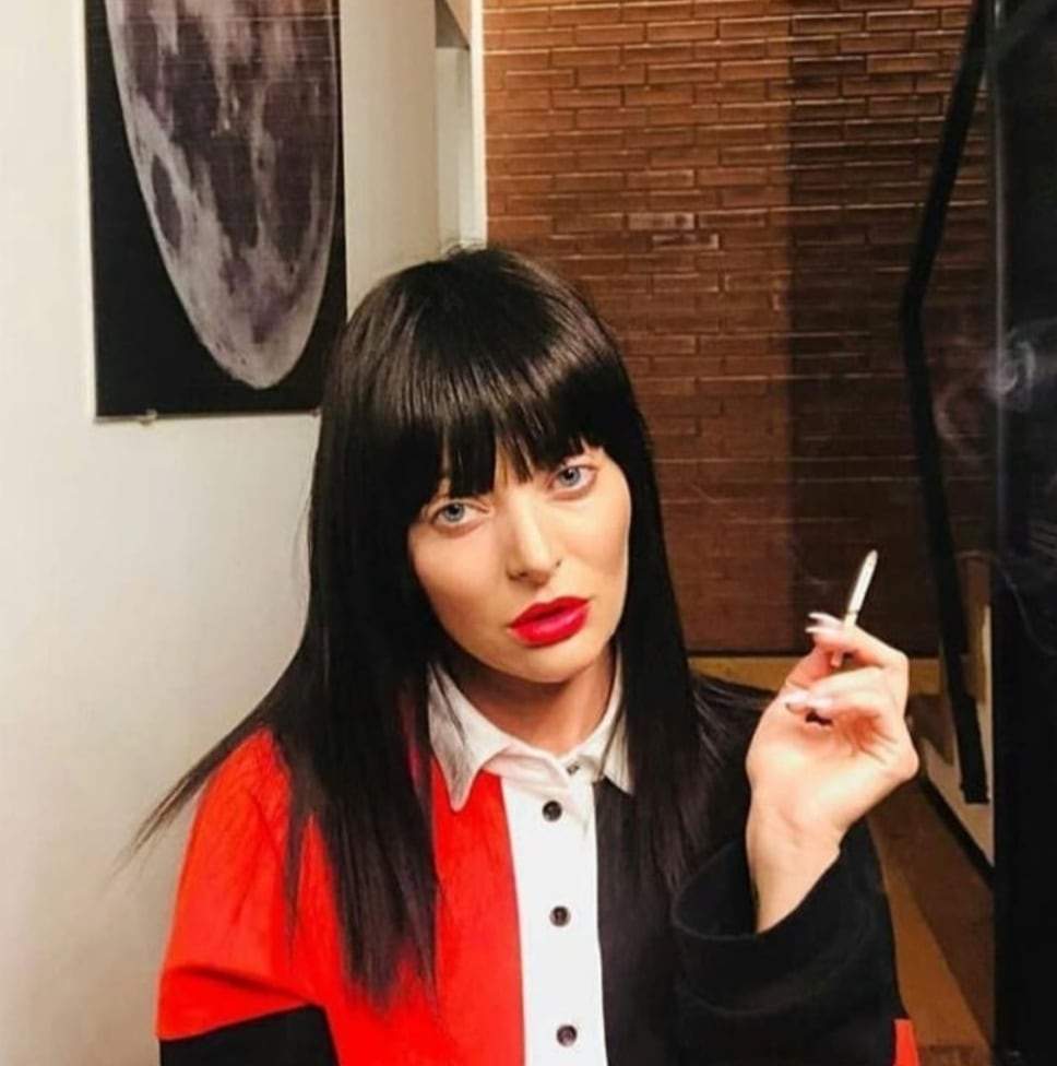 Delia, îmbrăcată în roșu și negru, brunetă, cu breton, cu o țigară în mână