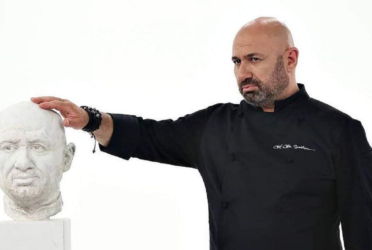 Cătălin Scărlătescu poartă o uniformă neagră de bucătar. Chef-ul ține mâna pe o statuie cu chipul său.