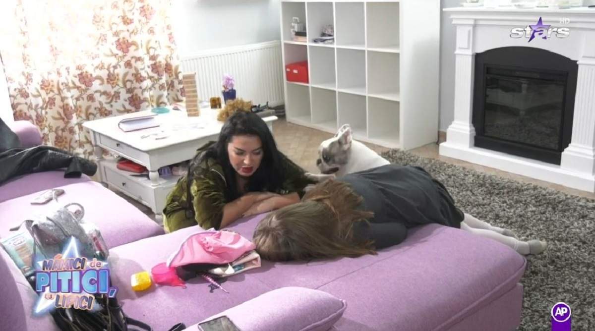 Fiica Oanei Roman și Elena Ionescu se află în dormitor. Isabela stă întinsă pe pat și poartă o rochiță gri, iar artista poartă un trening verde și-și ține brațele pe pat pentru a vorbi cu ea.