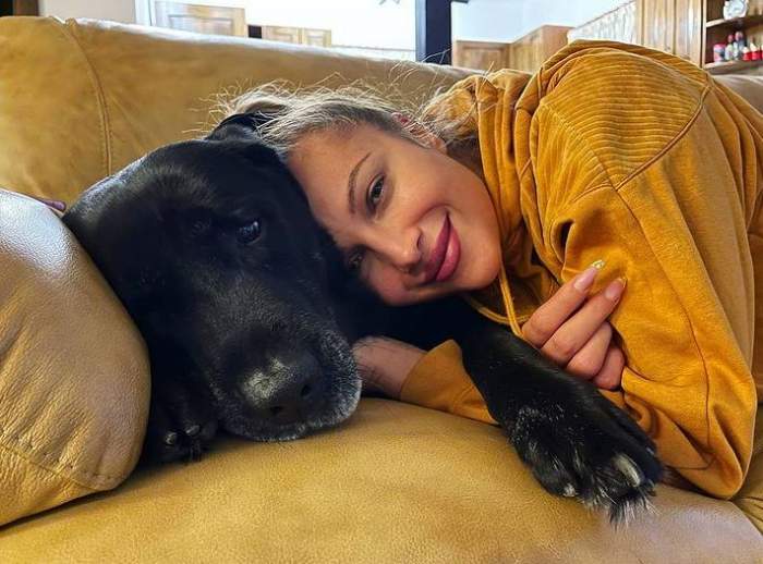Lora stă pe canapea cu câinele ei. Artista poartă un hanorac galben și zâmbește, ținându-și animalul în brațe.