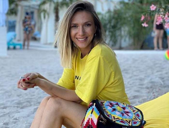Adela Popescu se află pe plajă. Artista poartă un tricou galben și zâmbește larg.