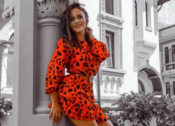 Cristina Șișcanu poartă o rochie oranj spre roșu, tip animal print. Vedeta zâmbește și se sprijină de un zid.