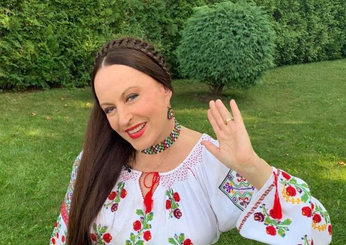 Maria Dragomiroiu e în parc. Vedeta poartă o ie albă cu flori roșii și face cu mâna.