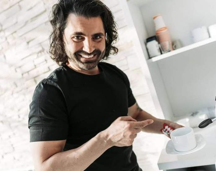 Pepe e în bucătărie. Artistul poartă un tricou negru și ține în mână o ceașcă de cafea.