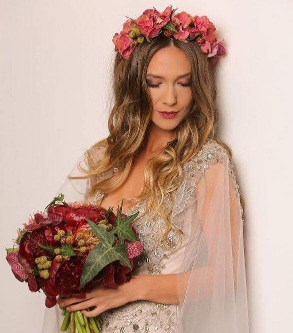Adela Popescu poartă o rochie albă cu brațe din voal și are pe cap o coroniță de flori roz. Vedeta privește în jos și ține în mână un buchet mare de flori roșii.