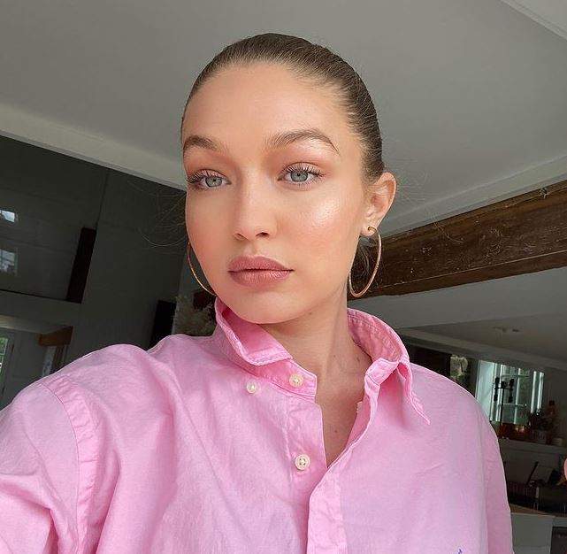 Gigi Hadid își face un selfie. Vedeta poartă o cămașă roz și cercei rotunzi aurii.