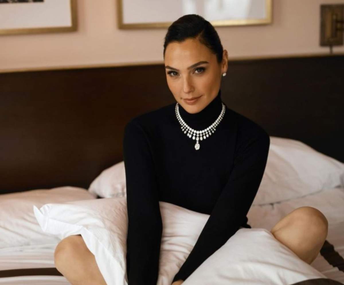 Gal Gadot este in pat, poarta o bluza cu guler ridicat neagra, un colier de diamante si are parul prins