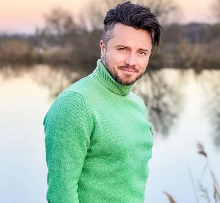 Tavi Clonda poartă un pulover verde și zâmbește larg. În spatele artistului se vede un lac.