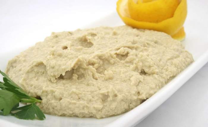Hummus așezat pe o farfurie albă. Lângă el sunt câteva frunze de pătrunjel și felii de lămâie.