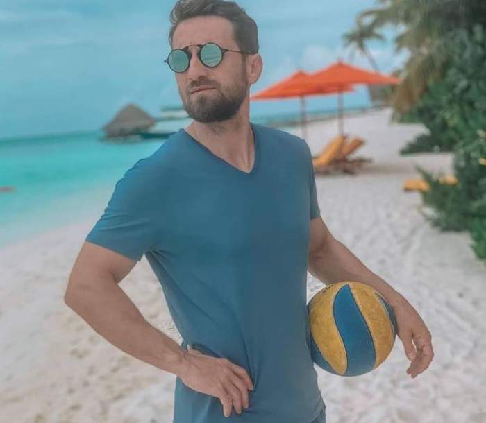 Dani Oțil e pe plajă. Prezentatorul poartă un tricou bleu și ochelari de soare, are o mână în șold, iar cu cealaltă ține o minge la nivelul bazinului, colorată în galben și bleu.