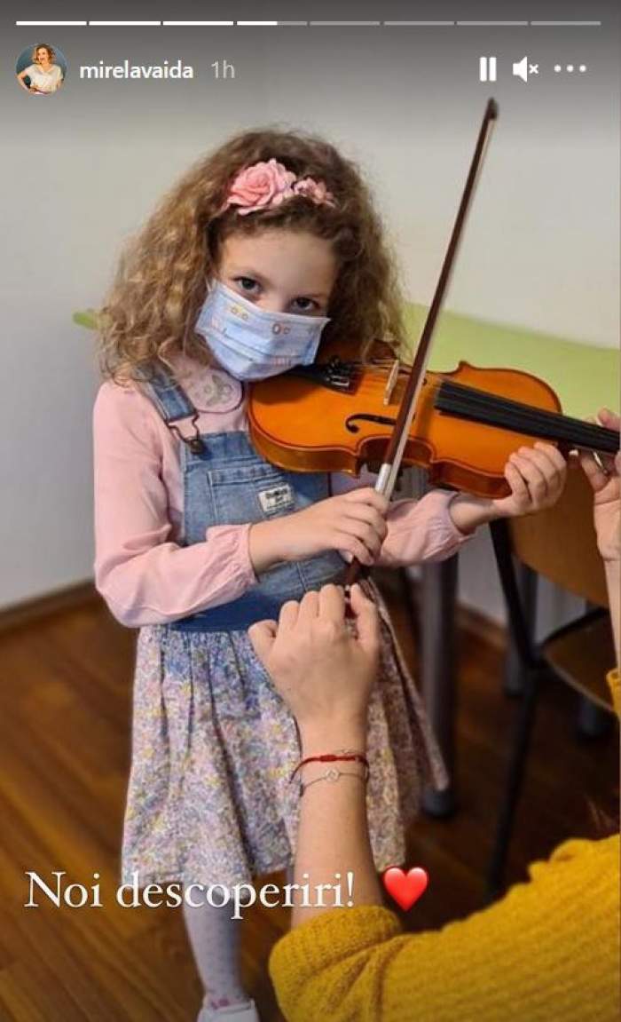 Fiica Mirelei Vaida e îmbrăcată într-o bluză roz și un sarafan din denim albastru. Carla poartă mască de protecție și cântă la vioară.