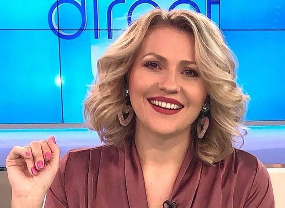 Mirela Vaida se află în platoul „Acces Direct”. Vedeta stă pe scaun și poartă o bluză roz închis spre mov. Artista zâmbește larg.