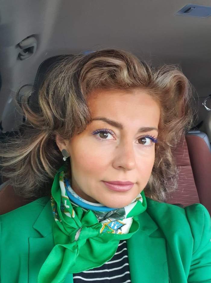 Alexandra Velniciuc e în mașină. Artista poartă o bluză neagră cu dungi albe, sacou verde și eșarfă verde.